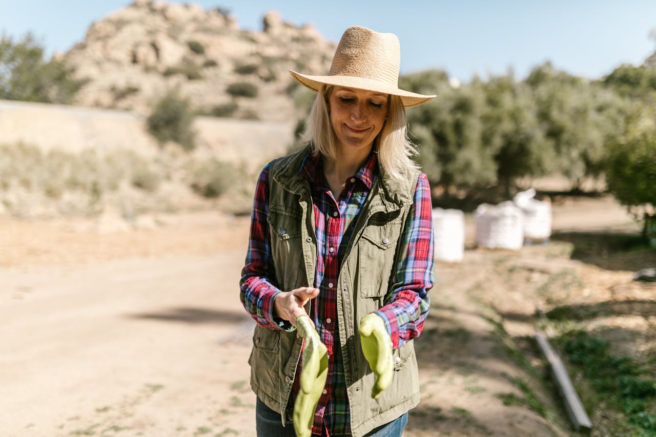 Woman farmer in a field wearing a vest and hat, showing rubber gloves.